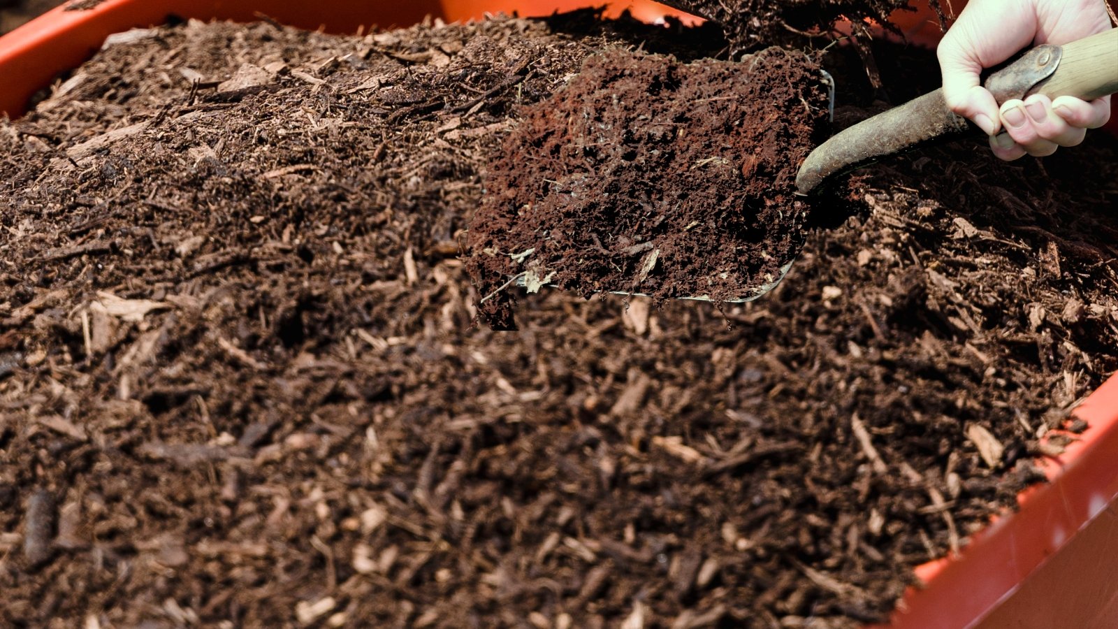 A gloved hand uses a small garden tool to mix dark, rich soil in a raised garden bed, revealing layers of decomposing organic material underneath.