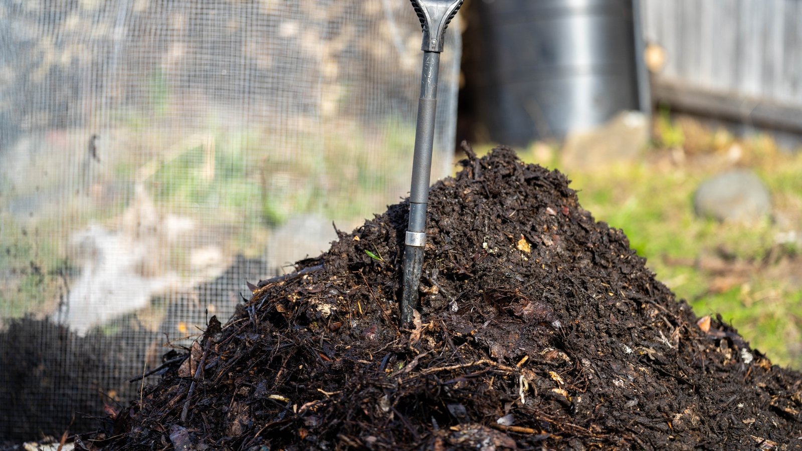 A large mound of rich dark earth sits in an outdoor space, surrounded by a wire fence, with a metal thermometer sticking out of the top to monitor the temperature.