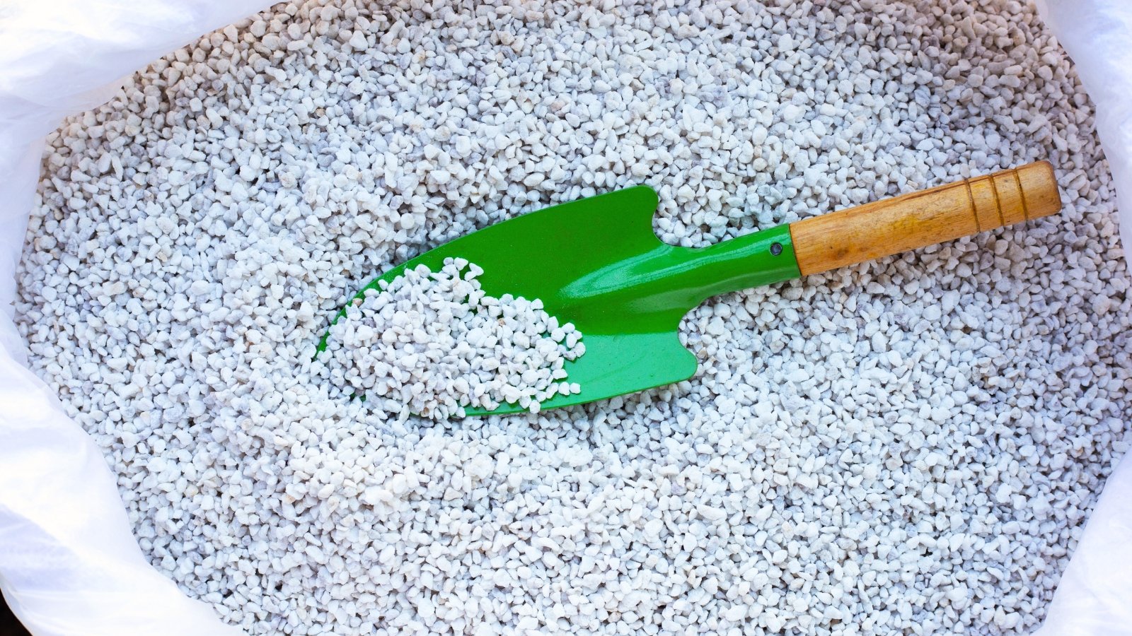 Small, lightweight, white, porous granules with a rough texture fill a white bag, accompanied by a green shovel placed inside.
